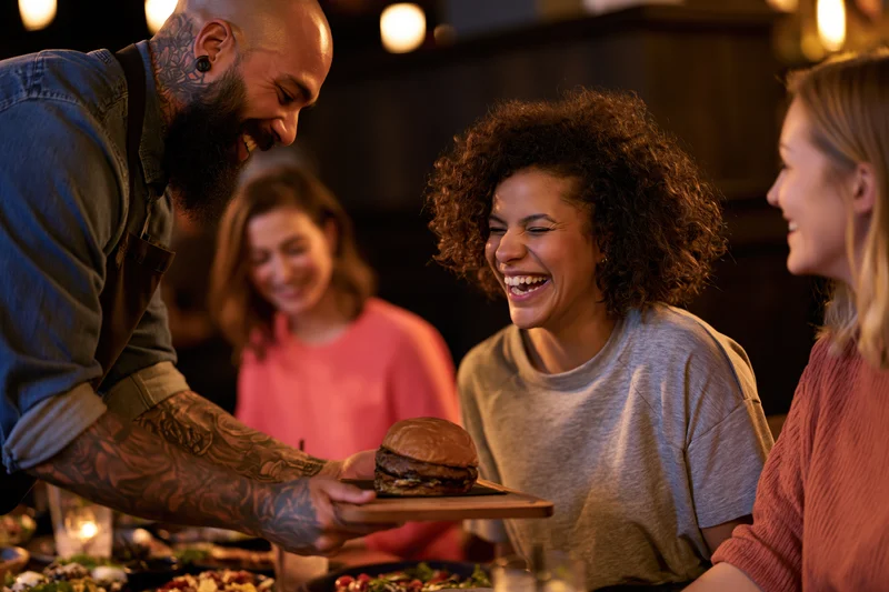 Tattooed waiter serving a burger to a laughing woman at a restaurant table