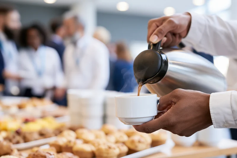 Hands pouring hot coffee from a steel thermal carafe into a white cup at a breakfast buffet with pastries and professionals in the background