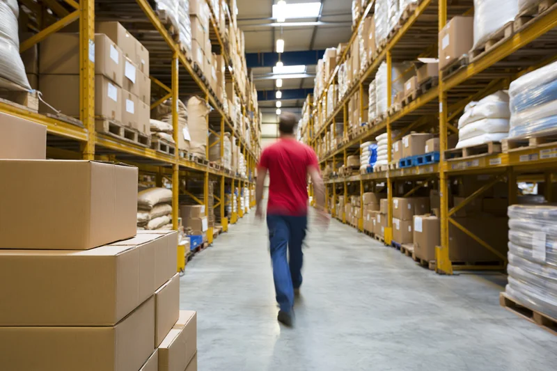 Man in red shirt and blue trousers walking through warehouse aisle between tall shelves of boxes and pallets
