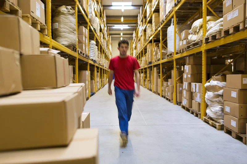 Man in red shirt and blue trousers walking through warehouse aisle between stacked boxes and pallets