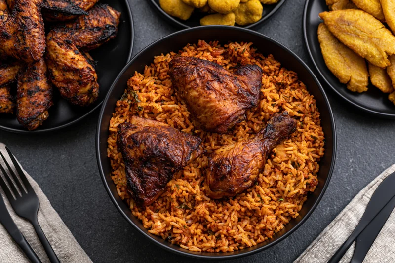 Jollof rice with roasted chicken pieces served in a black bowl surrounded by side dishes on a dark surface