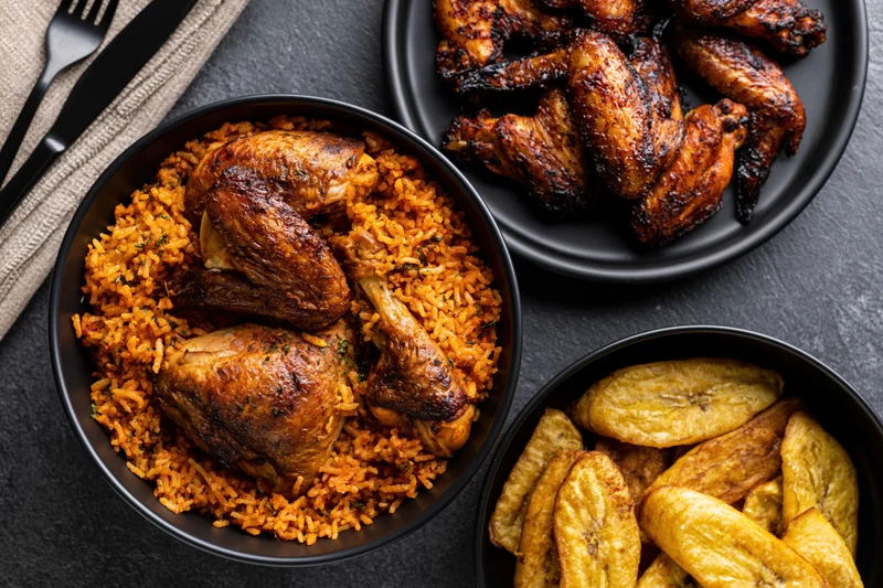 Jollof rice with roasted chicken pieces and fried plantains served in black bowls on a dark stone table