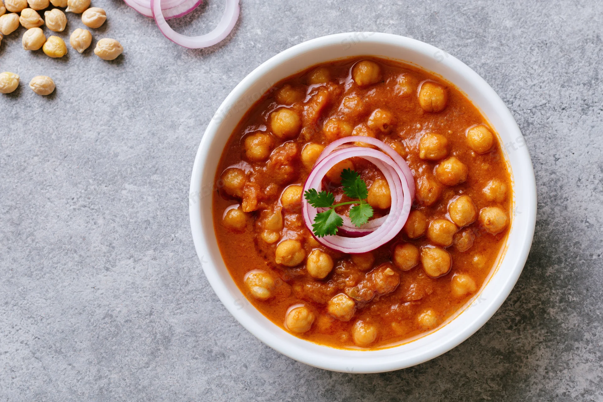 Chana masala in a white ceramic bowl topped with sliced onion and cilantro on a gray surface