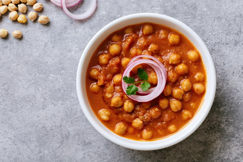 Chana masala in a white ceramic bowl topped with sliced onion and cilantro on a gray surface