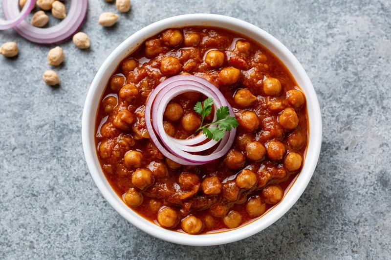 Chana masala in a white ceramic bowl garnished with red onion and cilantro on a gray stone surface
