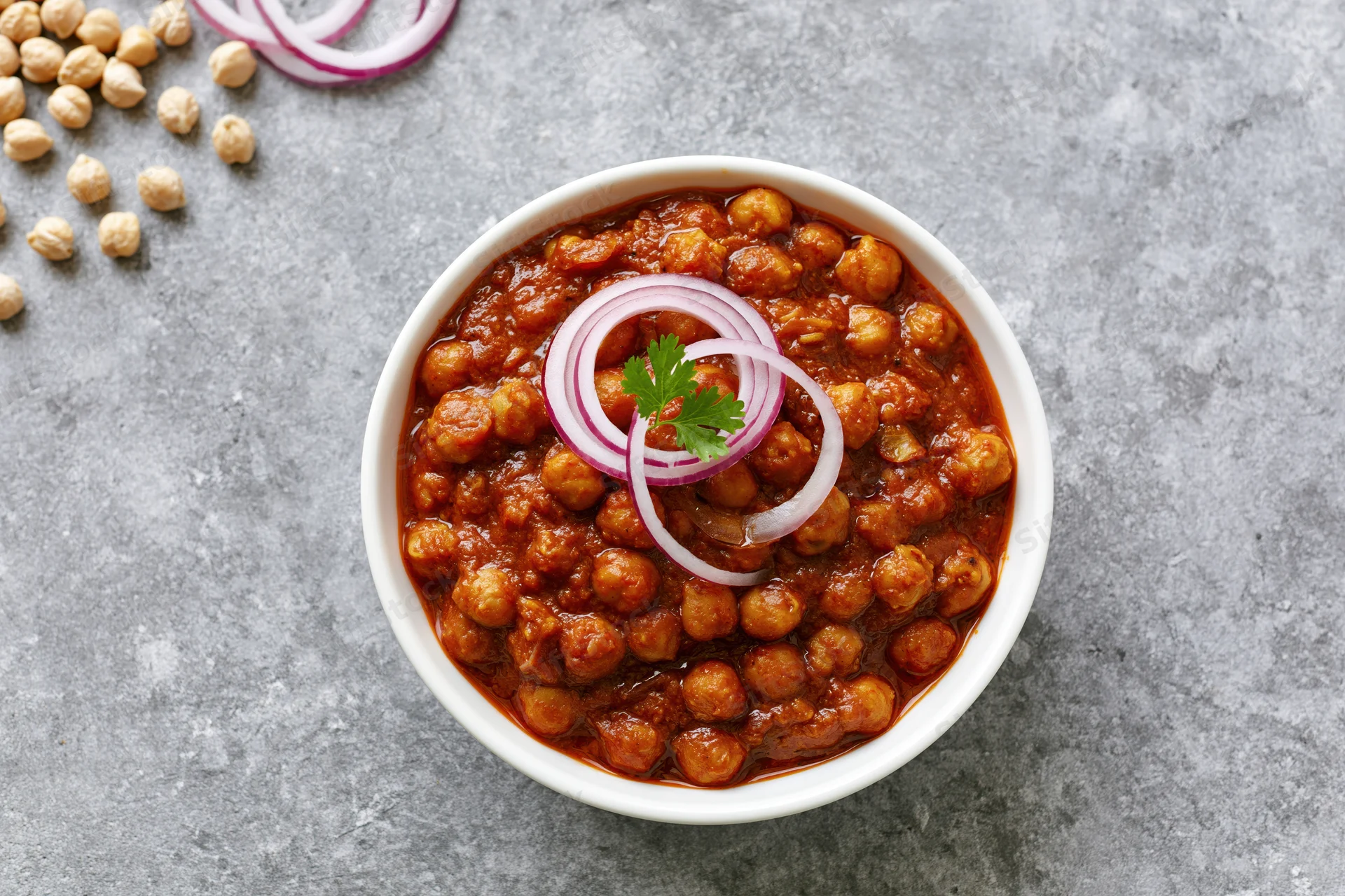 Chana masala in a white ceramic bowl garnished with sliced red onion and parsley on a gray stone surface