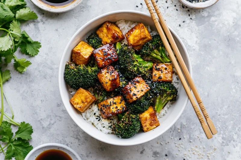 Teriyaki tofu and broccoli over rice in a white ceramic bowl with chopsticks and sesame seeds on a gray countertop