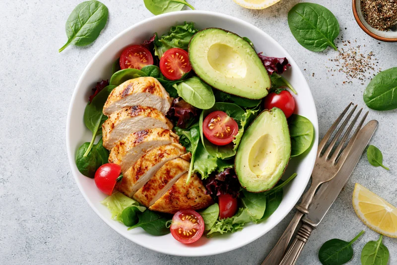 Grilled chicken and avocado salad in a white bowl with fresh greens and tomatoes on a gray surface