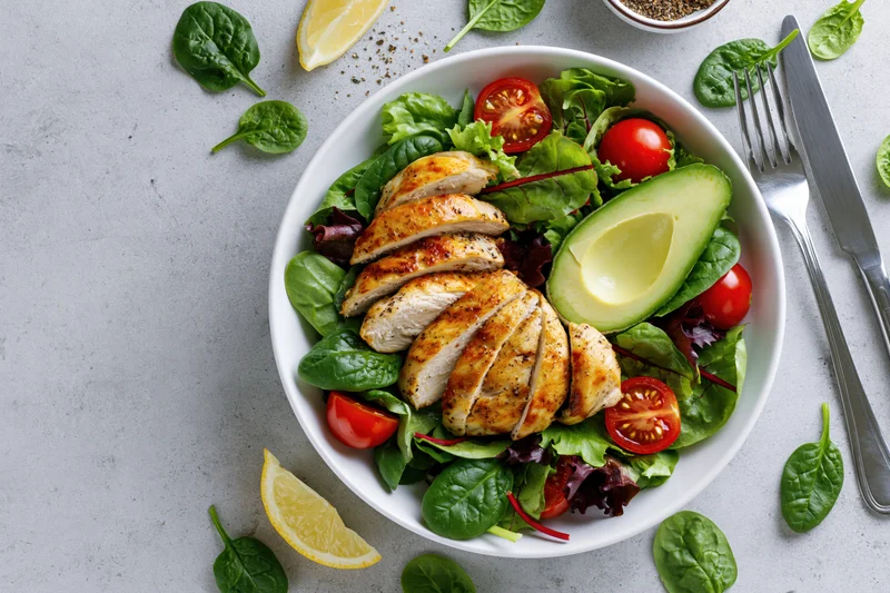 Grilled chicken salad with avocado and tomatoes in a white bowl on a gray surface