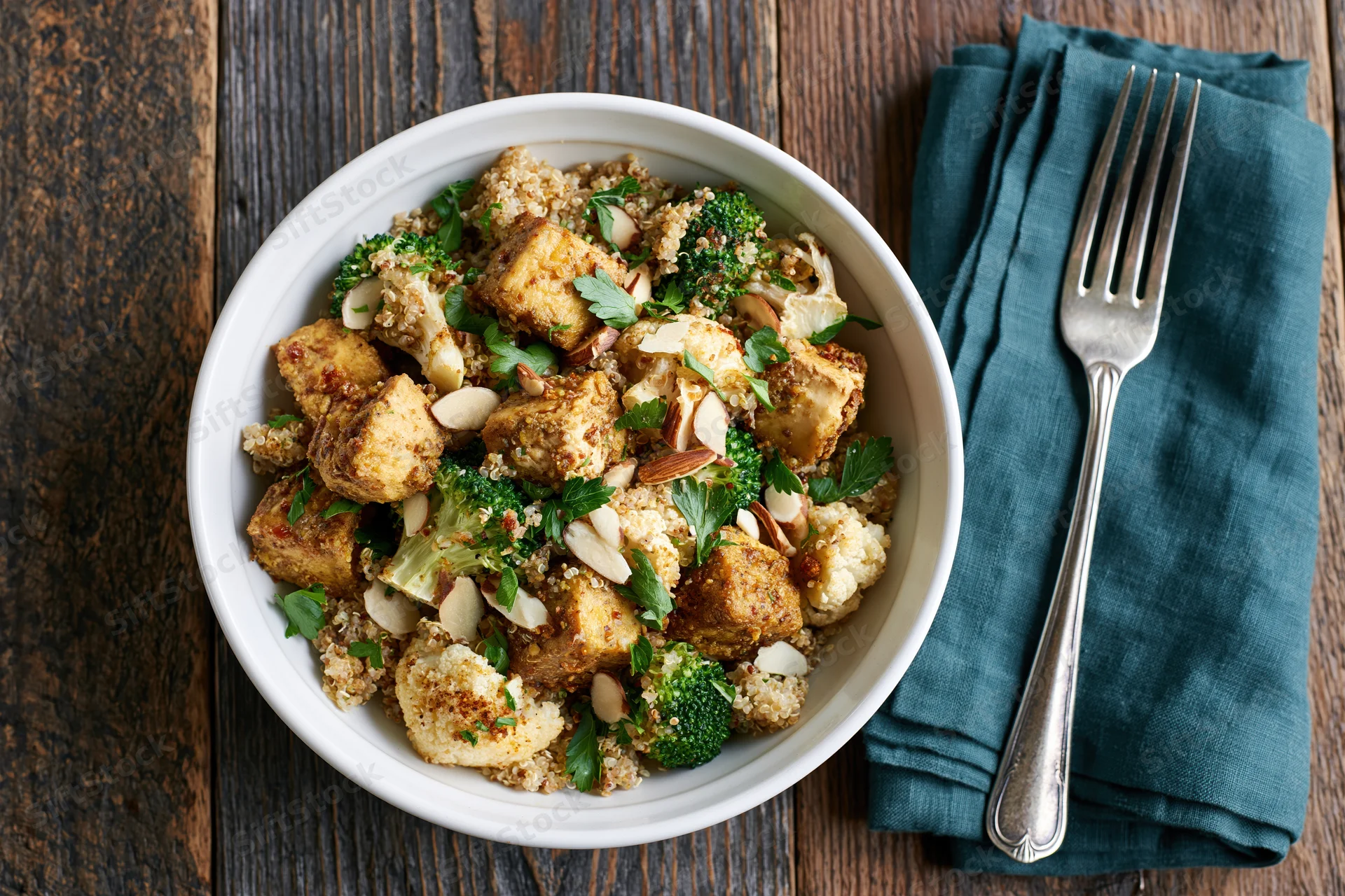 White ceramic bowl filled with spiced roasted tofu cubes, broccoli, cauliflower, quinoa, and almonds on wooden table with fork and teal napkin