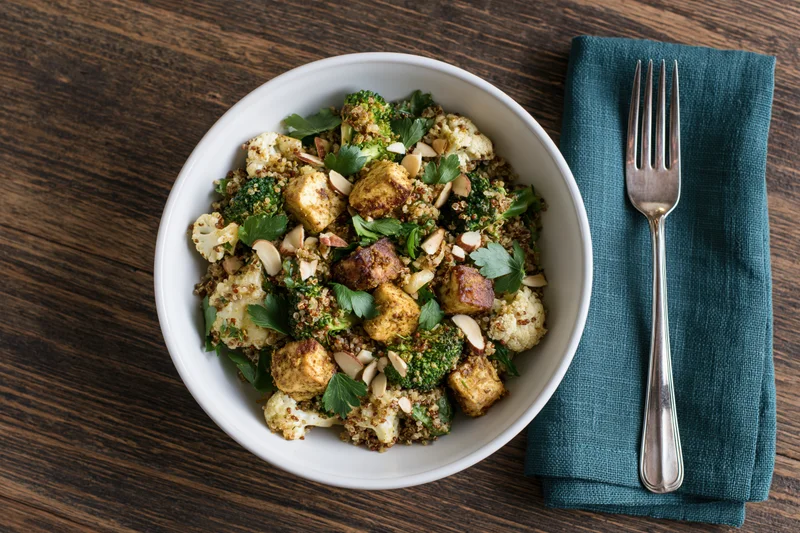 A bowl of spiced roasted tofu and vegetable salad with quinoa and almonds on a wooden table beside a fork on a teal napkin