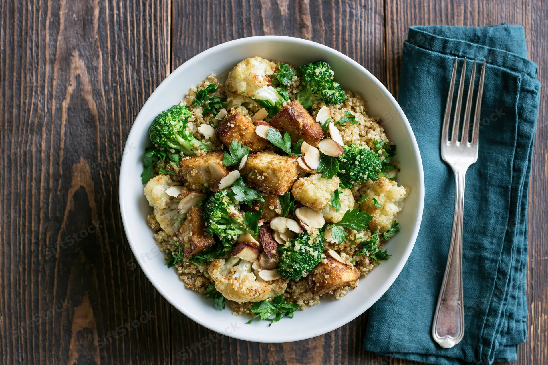 White ceramic bowl with spiced roasted tofu, broccoli, cauliflower, quinoa, and almonds on dark wooden table with fork and teal napkin