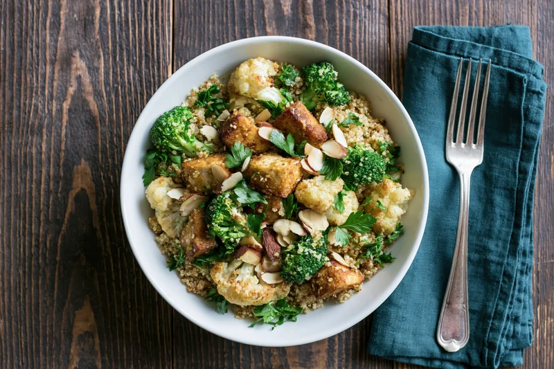 White ceramic bowl with spiced roasted tofu, broccoli, cauliflower, quinoa, and almonds on dark wooden table with fork and teal napkin