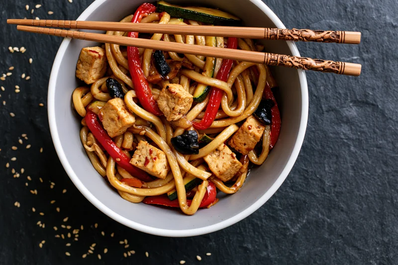 White ceramic bowl of stir-fried udon noodles with tofu cubes and vegetables, chopsticks resting on top, on dark slate surface