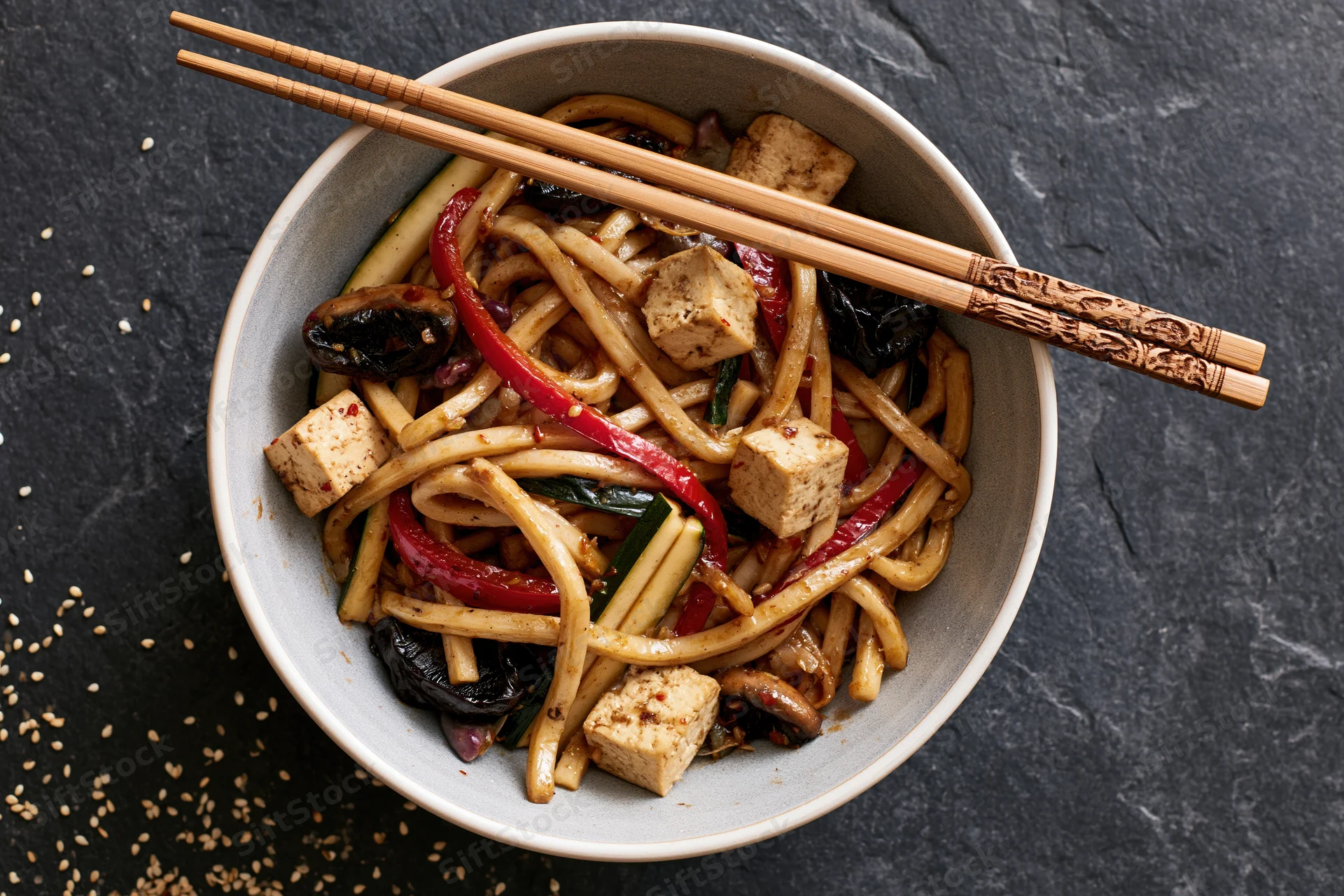 White ceramic bowl of stir-fried udon noodles with tofu and vegetables on dark slate surface