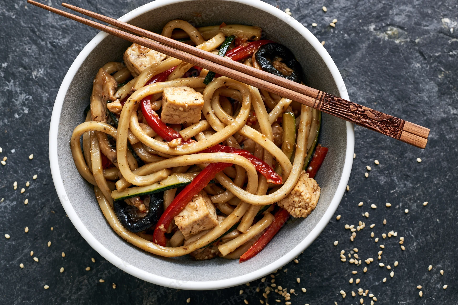 Udon noodles stir-fried with tofu cubes and vegetables in a white ceramic bowl with chopsticks on a dark stone surface