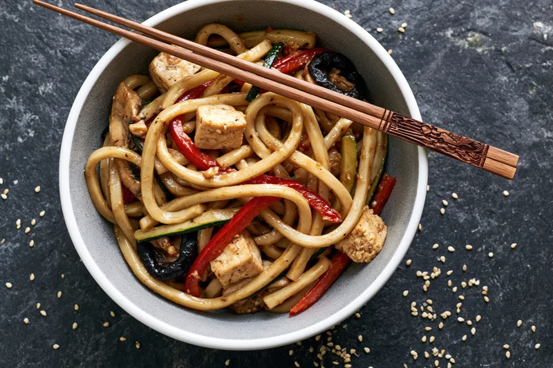 Udon noodles stir-fried with tofu cubes and vegetables in a white ceramic bowl with chopsticks on a dark stone surface