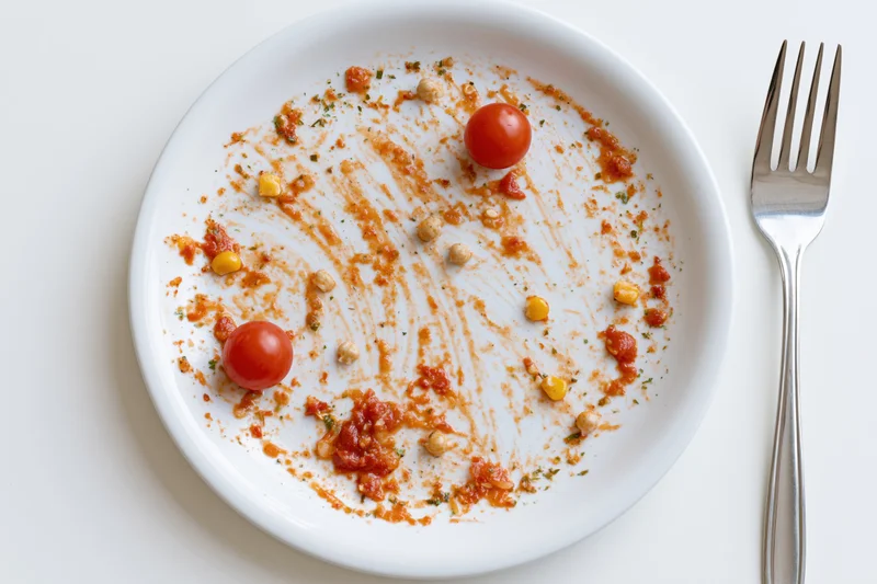 Empty white ceramic plate with tomato sauce residue and cherry tomatoes beside a fork on a white surface