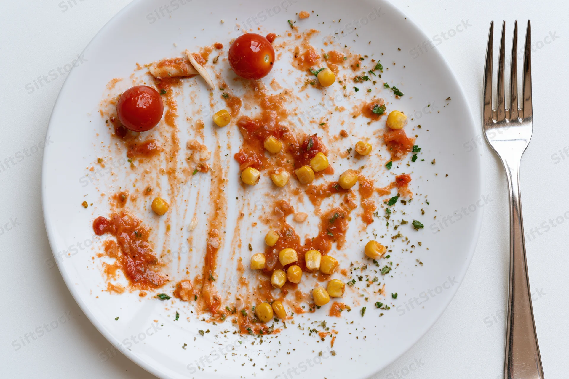 Empty white ceramic plate with tomato sauce residue and scattered cherry tomatoes and corn kernels beside a fork on a white surface