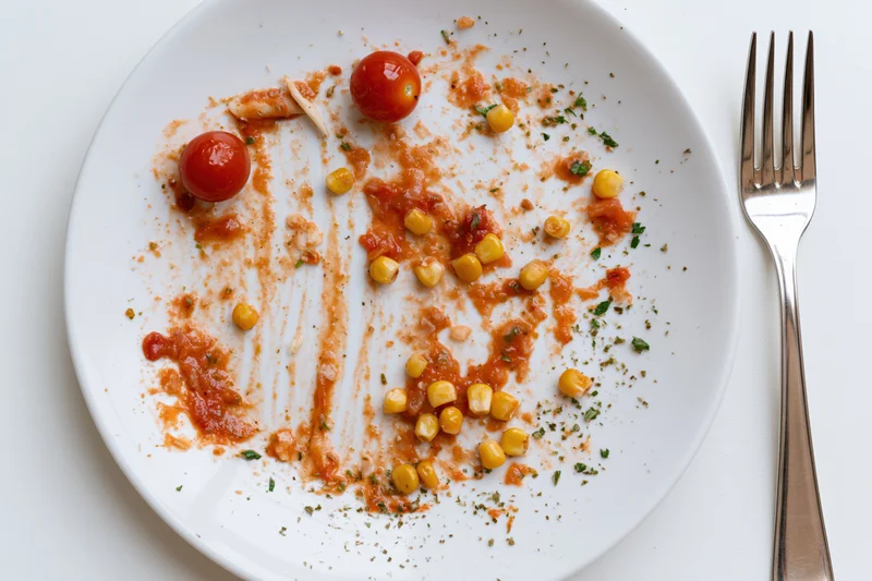 Empty white ceramic plate with tomato sauce residue and scattered cherry tomatoes and corn kernels beside a fork on a white surface