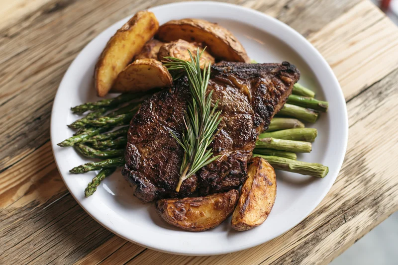 Seared ribeye steak with roasted potatoes and asparagus on a white plate on wooden table