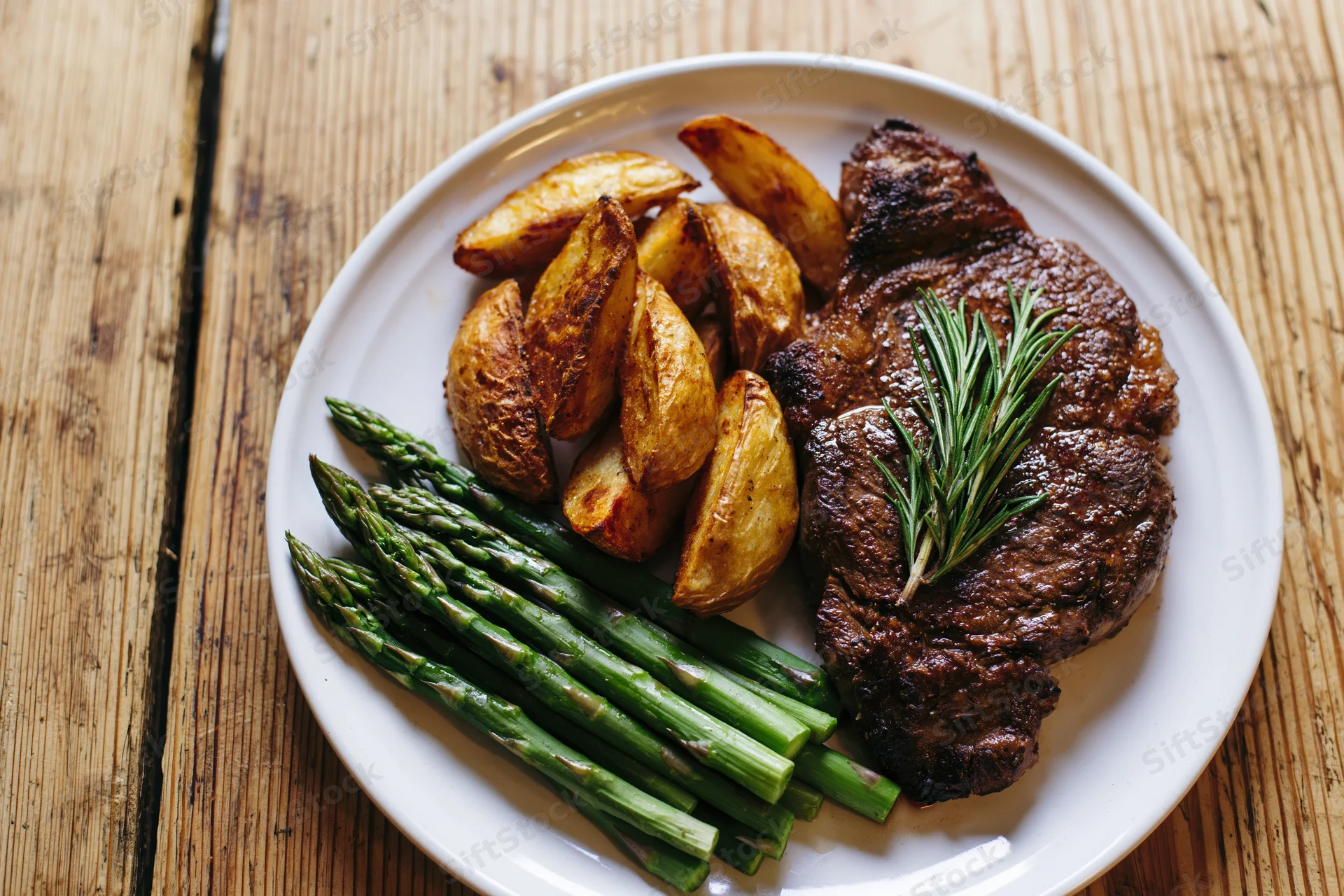 Seared ribeye steak with roasted potatoes and asparagus on a white plate on a wooden table