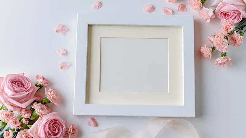 White square frame with blank cream mat surrounded by pink roses and carnations on a light surface with ribbon and petals
