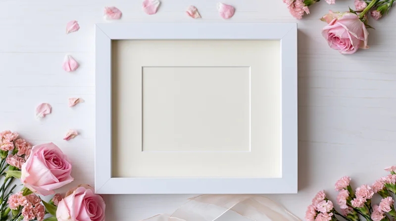 White frame with blank mat surrounded by pink roses and carnations on a wooden surface