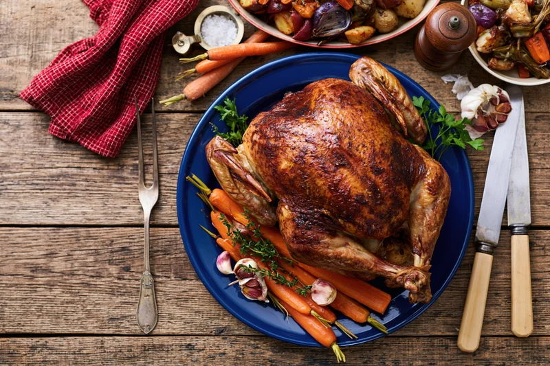 Whole roasted turkey on a blue enamel plate with carrots and herbs on a wooden table surrounded by dinnerware and seasonings