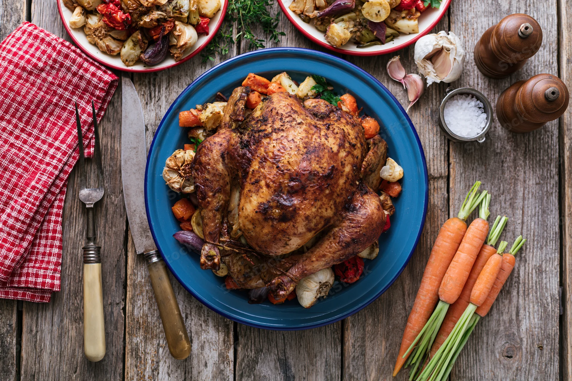 Whole roasted turkey on a blue enamel plate surrounded by vegetables and dinnerware on a rustic wooden table