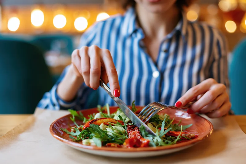 Woman eating grilled vegetable salad with fork and knife at restaurant table
