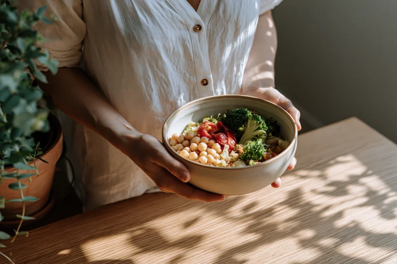 Woman holding a ceramic bowl of chickpeas broccoli and tomato salad on a wooden table