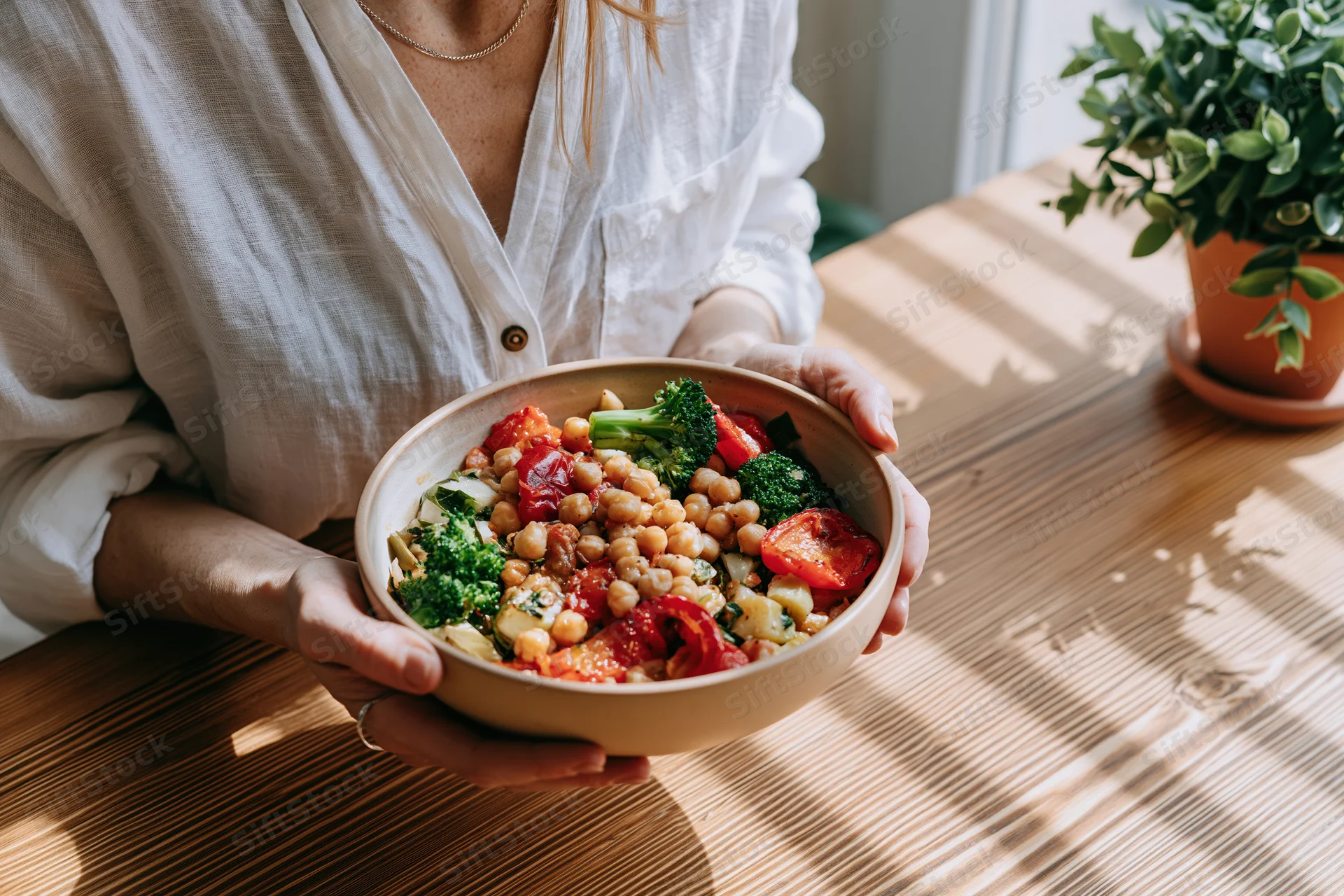 Woman holding a ceramic bowl filled with chickpeas, broccoli, and tomatoes on a wooden table