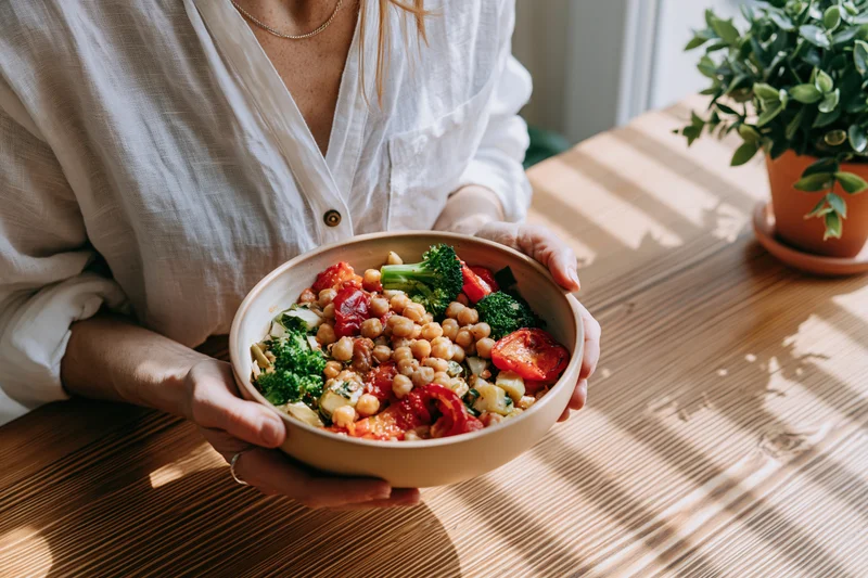 Woman holding a ceramic bowl filled with chickpeas, broccoli, and tomatoes on a wooden table