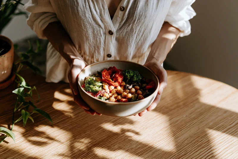Woman holding a ceramic bowl of chickpea salad with broccoli and tomatoes on a wooden table