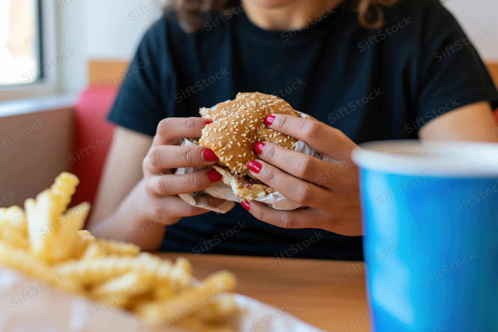 Woman holding a partially eaten sesame-seed burger with both hands at a diner table with fries and a drink in front of her
