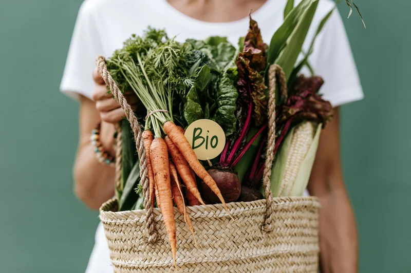 Woman holding a woven straw tote bag filled with fresh organic vegetables against a green background