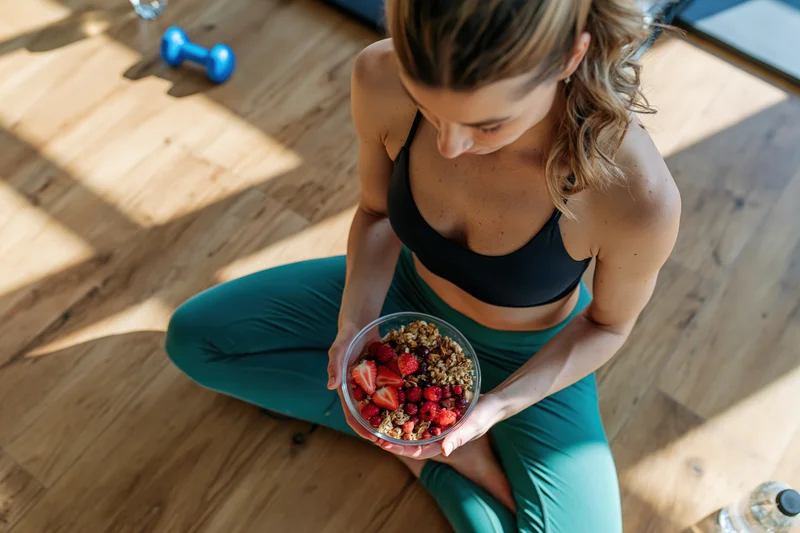 Woman in teal leggings and black sports bra sitting cross-legged on wooden floor holding a bowl of granola and berries after workout