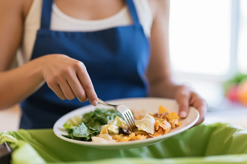 Woman scraping food scraps from a plate into a compost bin in a kitchen