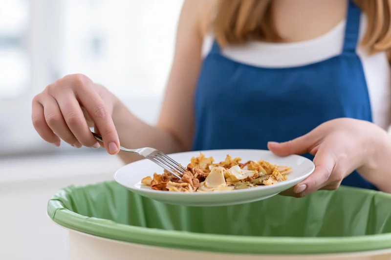 Woman scraping leftover food scraps from a white plate into a green compost bin