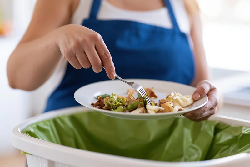 Woman scraping food scraps from a plate into a compost bin in a kitchen