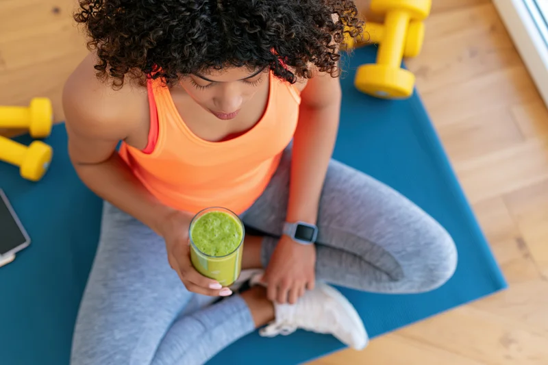 Woman sitting on blue yoga mat holding green smoothie after workout