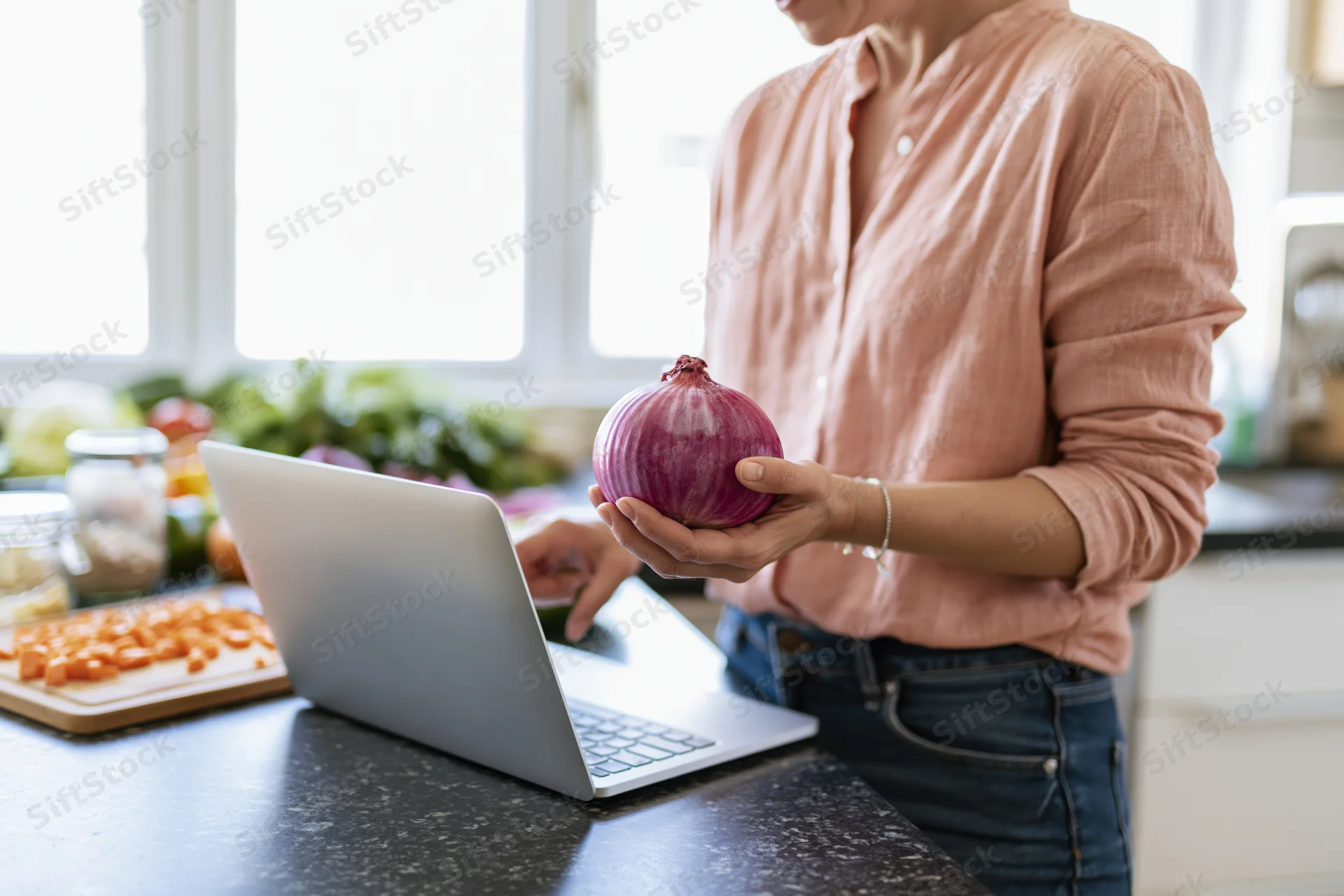 Woman holding red onion while using laptop on kitchen counter