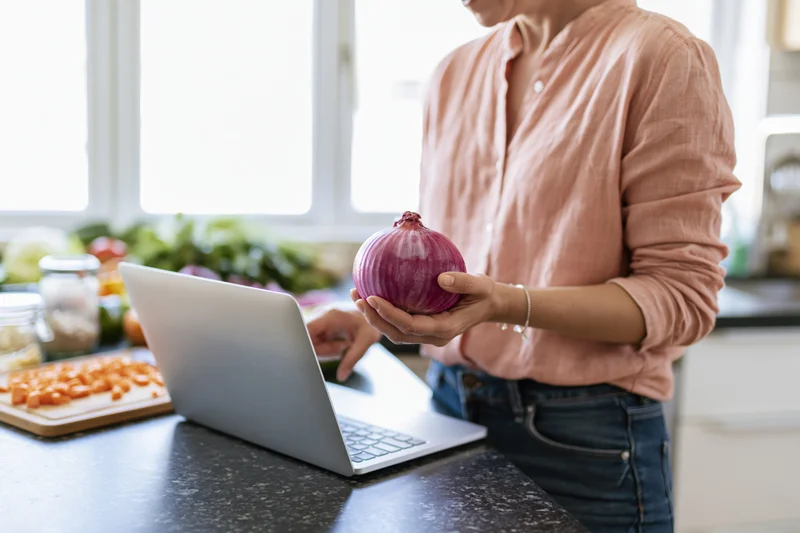 Woman holding red onion while using laptop on kitchen counter