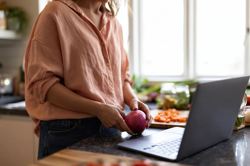 Woman holding red onion at kitchen counter beside open laptop