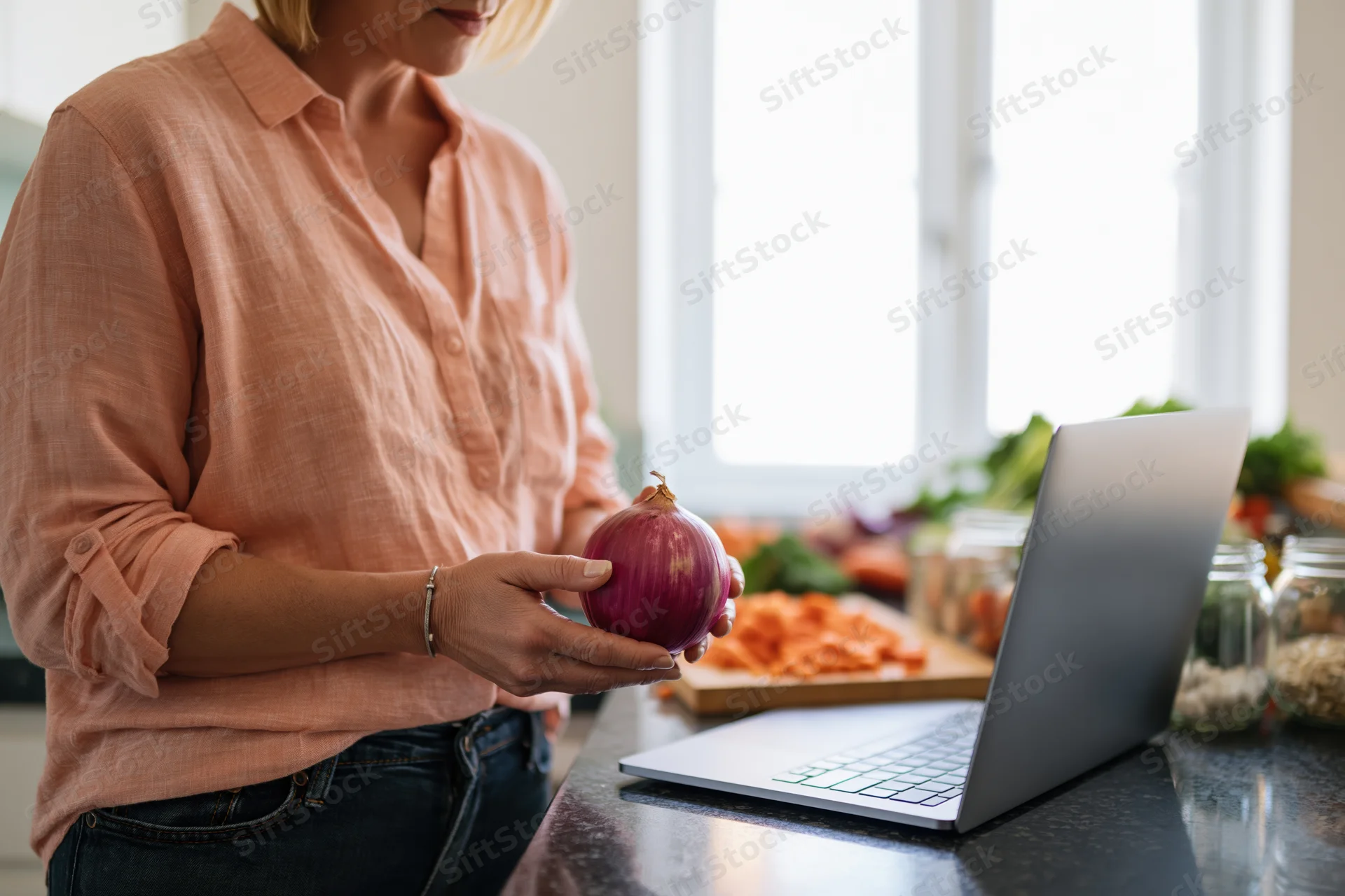 Woman holding red onion at kitchen counter with laptop and chopped vegetables nearby