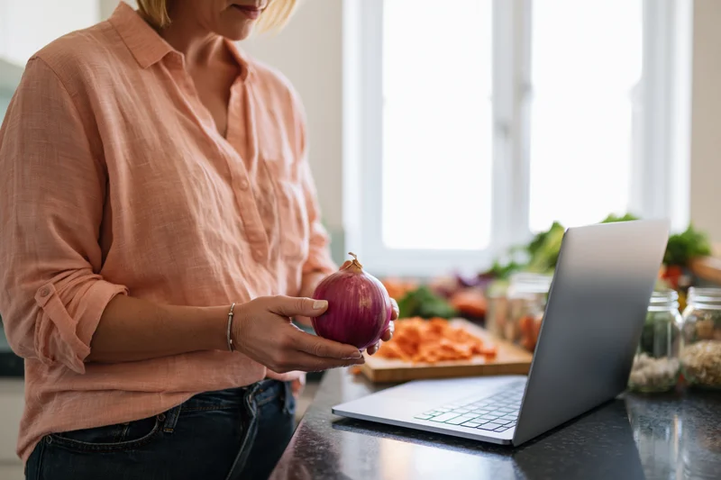 Woman holding red onion at kitchen counter with laptop and chopped vegetables nearby