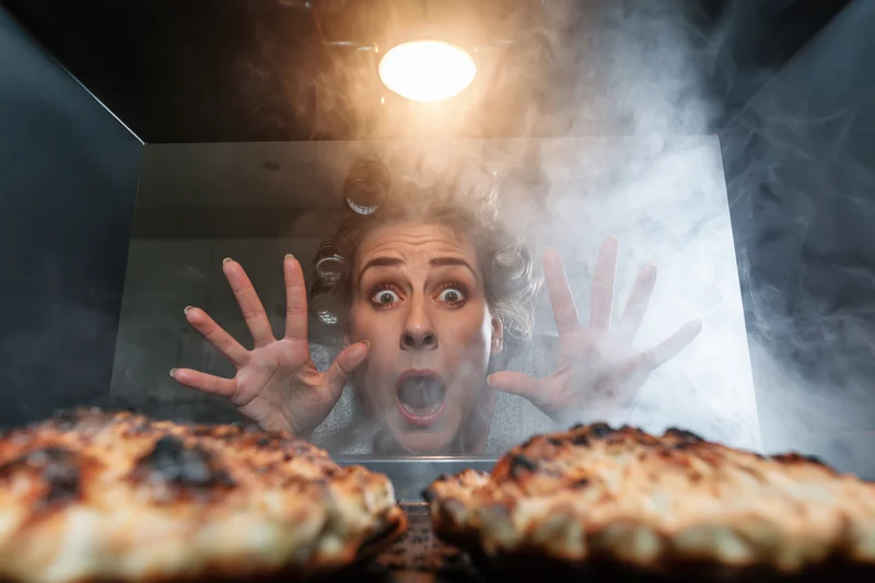 Woman with hair curlers reacting in horror with hands raised behind oven glass as burnt food smokes inside
