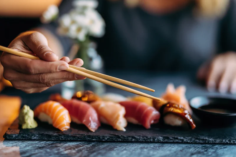 Woman's hand holding wooden chopsticks reaching toward a sushi platter on a slate plate