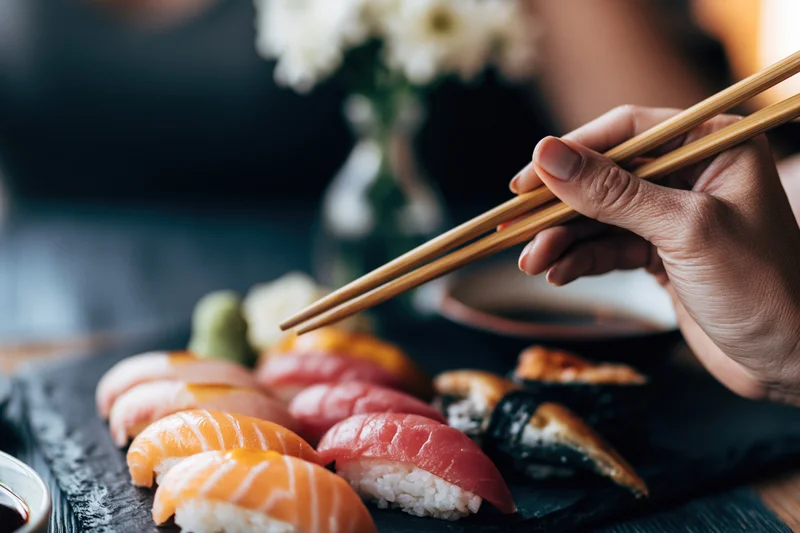 Woman's hand holding wooden chopsticks reaching toward sushi on a black stone plate
